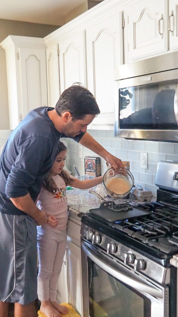 Dad and daughter making Kodiak Cake pancakes in a white kitchen at the stove.